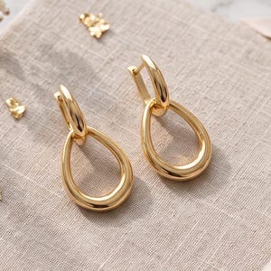 May include: A pair of gold-toned hoop earrings featuring a polished, teardrop-shaped pendant suspended from a circular hoop closure. The earrings have a smooth, reflective metallic finish and a simple, continuous loop design for the drop component.
