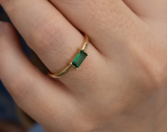 May include: A gold ring with a rectangular emerald green gemstone. The ring is on a person's finger.