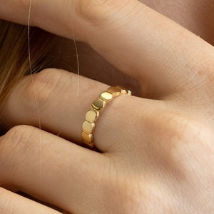May include: A gold ring with a repeating geometric design of small, connected, rounded rectangles. The ring is worn on a finger, with the hand and part of the arm visible. The ring is the focal point, showcasing a simple yet elegant jewelry piece.