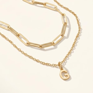 May include: Two gold-colored necklaces. The top necklace has a chain of elongated links. The bottom necklace has a delicate chain with a pendant of the letter "G". The necklaces are displayed on a white surface.