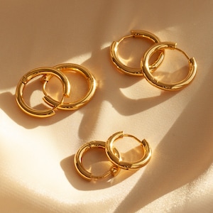 May include: Four gold hoop earrings of different sizes on a light beige fabric background.