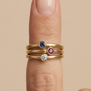 May include: Three gold rings stacked on a finger, each with a different gemstone. The top ring has a blue gemstone, the middle ring has a red gemstone, and the bottom ring has a clear gemstone. The rings are simple bands with the gemstones set in the center.