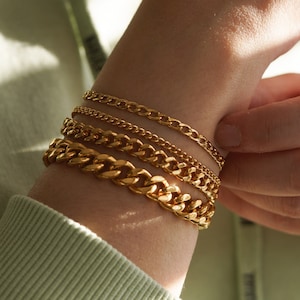 May include: A person wearing a stack of four gold chain bracelets on their wrist. The bracelets are different widths and styles, creating a layered look.