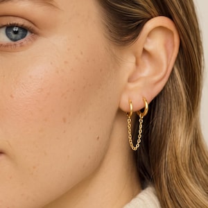 May include: A pair of gold hoop earrings connected by a delicate gold chain. The two small hoops are worn in adjacent piercings on the earlobe, with the fine link chain draping between them to create a connected, layered appearance in polished gold.