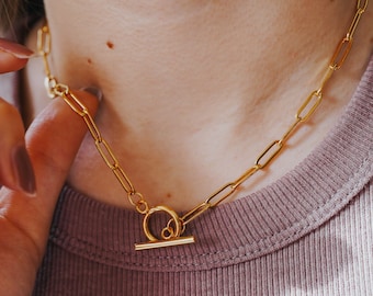 May include: A gold-colored paperclip toggle necklace. The necklace is made of elongated rectangular links and features a circular toggle clasp. The text "Tiny Paperclip Toggle Necklace" is visible at the bottom of the image.