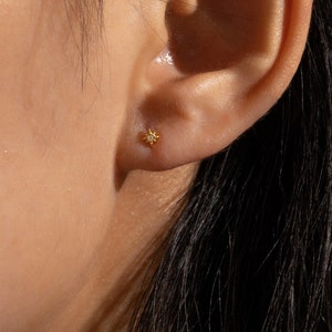 May include: A close-up of a person's ear with a small, gold-colored star-shaped stud earring. The earring is a simple design, with a small, raised center and pointed edges. The skin around the ear is smooth and has a warm tone.