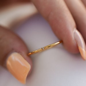 May include: A delicate gold ring with a small, clear gemstone set in the center. The ring is thin and minimalist in design, with a polished finish. The ring is held between fingers with a light peach-colored nail polish.