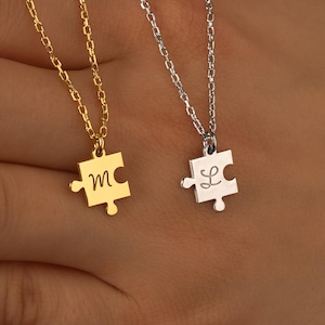 May include: Two delicate necklaces with puzzle piece pendants. One is gold-toned with the letter "m", the other is silver-toned with the letter "L". Both pendants hang from fine chains. The necklaces are displayed against a neutral background.