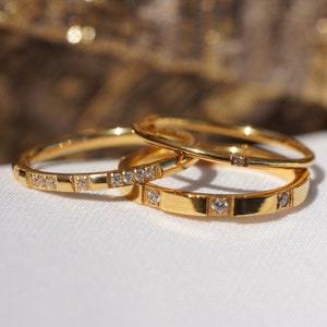 May include: Three gold rings with small, clear gemstones set into the bands. The rings are stacked on top of each other, with the focus on the bottom ring. The rings are on a white surface with a blurred gold background.