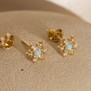 May include: A pair of gold star-shaped earrings with a white opal stone in the center of each star. The stars are surrounded by small clear stones.
