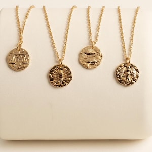May include: Four gold-toned necklaces with circular pendants, each featuring a distinct embossed zodiac symbol. The pendants hang from delicate matching gold chains, displaying detailed engravings of Libra, Scorpio, Pisces, and Virgo signs.