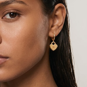 May include: A gold hoop earring featuring a small, heart-shaped pendant charm. The heart charm is engraved with the capital letter S in the center. The earring is made of a polished gold-tone metal and hangs from a circular hoop closure.