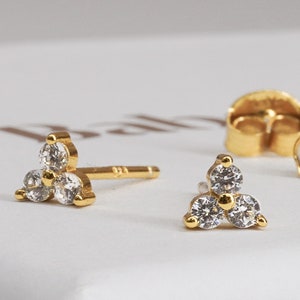 May include: A pair of gold stud earrings with three small clear cubic zirconia stones set in a triangular shape.