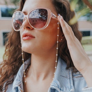 May include: Oversized, light pink sunglasses with a gold chain eyeglass holder featuring small white pearls. The sunglasses have a light pink tint and are worn with a light blue denim jacket.