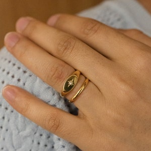 May include: A gold-colored ring set. One ring features a black oval with a starburst design and a small diamond. The other ring is a simple, smooth band. The rings are worn on a hand.