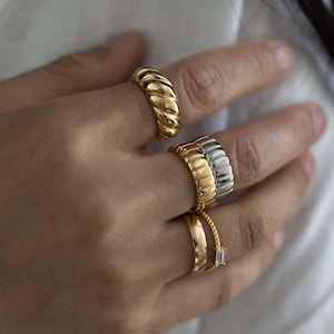 K&ouml;nnte beinhalten: Nahaufnahme einer Hand mit mehreren goldenen und silbernen Ringen. Die Ringe haben verschiedene Designs, darunter ein dickes, geriffeltes Band, ein verdrehter Seil-Stil und ein Band mit einem rechteckigen Edelstein.