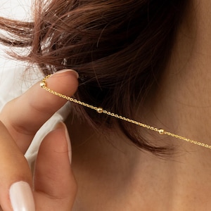 May include: A delicate gold necklace featuring small, evenly spaced beads. The necklace is held up against a person's skin, showcasing its fine chain and minimalist design. The gold color provides a warm contrast against the skin tone.