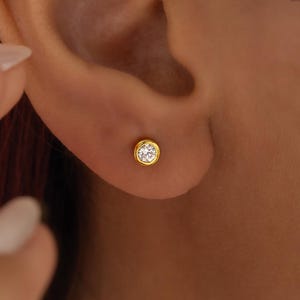 May include: A close-up of a gold stud earring with a clear, round gemstone. The earring is set in a gold bezel and is worn on an ear. The skin tone is light brown.