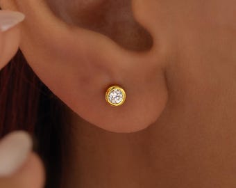 May include: A close-up of a gold stud earring with a clear, round gemstone. The earring is set in a gold bezel and is worn on an ear. The skin tone is light brown.