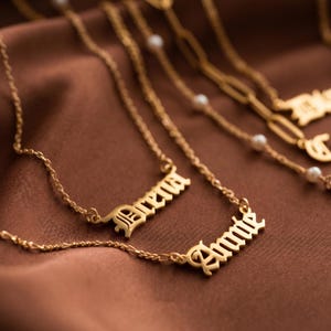 May include: Gold necklaces with name pendants, displayed on a brown satin surface. One necklace spells "Ariana" in an old English font, and another says "Annie". Other necklaces with pearl accents are visible in the background.