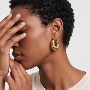 May include: Gold-colored, textured hoop earrings. The earrings have a ridged design and are worn on the ear. The image shows a person with short, dark hair and a cream-colored top.