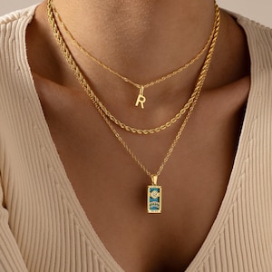 May include: A layered gold necklace set. The top chain has a small letter "R" pendant. The middle chain is a rope style. The bottom chain has a rectangular pendant with a blue and gold design. The necklaces are worn on a person wearing a cream-colored top.