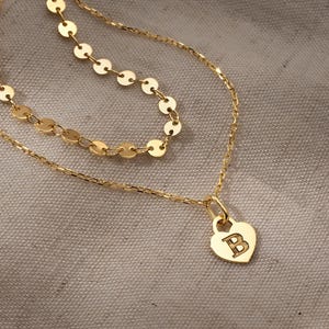 May include: A layered gold necklace with a heart pendant. The pendant features the letter "B". The necklace has two strands, one with small, round charms and the other with a delicate chain. The jewelry is displayed on a neutral background.