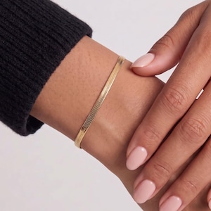 May include: A gold-toned herringbone chain bracelet with a flat, flexible link design. The bracelet features a polished, reflective surface that catches the light, creating a smooth, continuous metallic band around the wrist area.