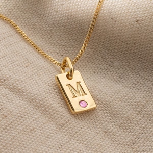 May include: A gold necklace with a rectangular pendant featuring the letter "M" and a small pink gemstone.