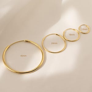 May include: Four gold-colored hoop earrings of varying sizes, ranging from 12mm to 40mm in diameter, are displayed. The earrings are arranged in a diagonal line, showcasing their different dimensions. The background is a soft, neutral color.