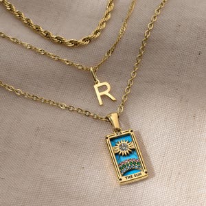 May include: Gold-toned layered necklaces. The top chain is a rope style, the middle has an "R" pendant, and the bottom features a rectangular pendant with a sun design and the text "THE SUN".