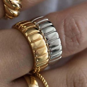 May include: Close-up of multiple gold and silver rings on a hand. The rings feature various designs, including a twisted rope, a ribbed band, and a smooth, curved band with a small, rectangular gemstone.