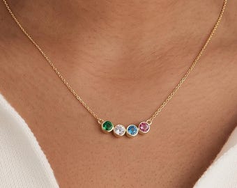 May include: A gold chain necklace featuring four small, round, bezel-set gemstones in a horizontal row. The stones are colored green, clear, blue, and pink. The necklace is worn around the neck, accompanied by small gold hoop earrings.