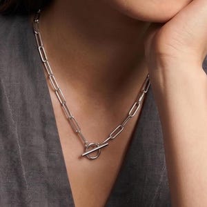 May include: A silver chain necklace with a toggle clasp is displayed. The necklace is paired with matching silver hoop earrings. The model is wearing a gray top. The jewelry is simple and elegant.