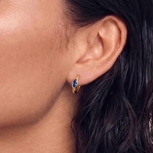 May include: A close-up of a person's face, with dark hair and gold hoop earrings. The earrings have a small, oval-shaped blue stone in the center. The skin tone is light brown, and the background is a soft pink.