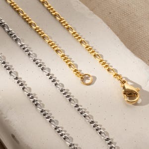K&ouml;nnte beinhalten: Zwei goldene Ketten, eine silberne Kette, alle im Curb-Link-Stil. Die goldenen Ketten haben einen Karabinerverschluss.