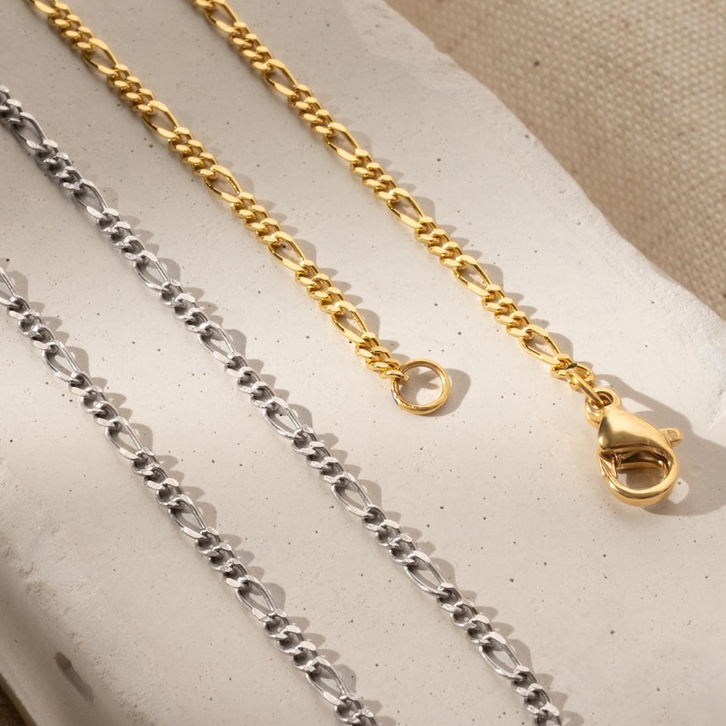 K&ouml;nnte beinhalten: Zwei goldene Ketten, eine silberne Kette, alle im Curb-Link-Stil. Die goldenen Ketten haben einen Karabinerverschluss.
