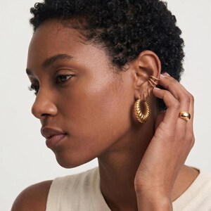 May include: A person wearing gold-coloured jewellery, including hoop earrings, a ring, and ear cuffs. The earrings have a textured, ridged design. The ring is a simple, rounded band. The ear cuffs have a twisted rope design. The jewellery is all gold-toned.