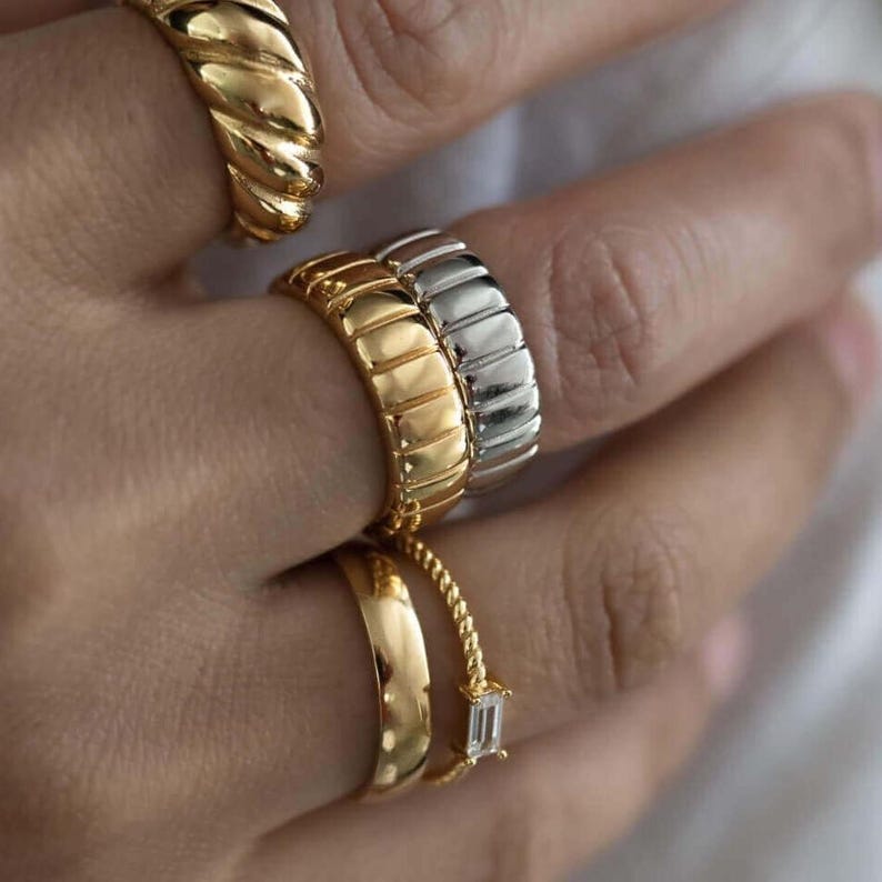 May include: Close-up of multiple gold and silver rings on a hand. The rings feature various designs, including a twisted rope, a ribbed band, and a smooth, curved band with a small, rectangular gemstone.