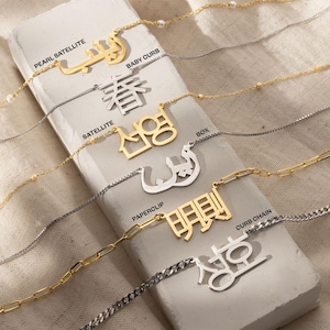 May include: A close-up of various gold and silver necklaces with different chain styles and personalized nameplates. The nameplates feature different languages and characters, including Arabic, Chinese, and Korean. The necklaces are displayed on a light brown textured surface.