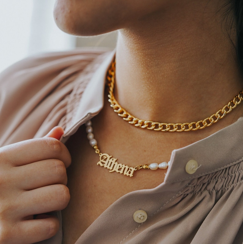 K&ouml;nnte beinhalten: Eine goldene Kettenkette mit einem goldenen Namensschild-Anh&auml;nger, auf dem "Athena" steht, und Perlenakzenten. Die Kette ist &uuml;ber eine dickere goldene Kette gelegt.