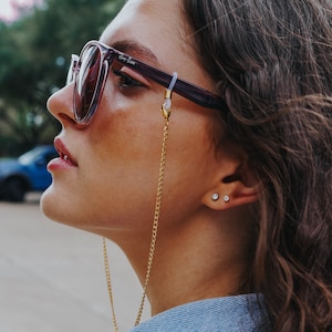 May include: A woman wearing purple-framed sunglasses with a gold chain eyeglass holder. The sunglasses have a Ray-Ban logo on the lens. The woman is also wearing small stud earrings.