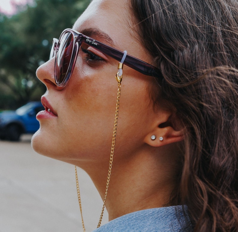 May include: A woman wearing purple-framed sunglasses with a gold chain eyeglass holder. The sunglasses have a Ray-Ban logo on the lens. The woman is also wearing small stud earrings.