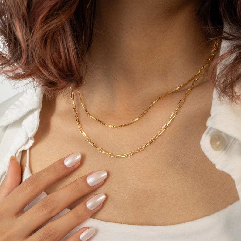 Puede incluir: Se muestran dos collares dorados. El collar superior es una fina cadena de caja, y el collar inferior es una cadena de clip. Los collares son usados por una persona que lleva una camisa blanca.