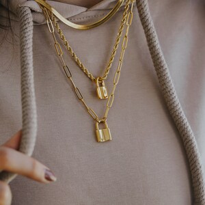 May include: Three gold necklaces with padlock pendants. The necklaces are layered on a person's neck. The top necklace is a thin, flat chain. The middle necklace is a thicker, rope chain. The bottom necklace is a chain with small, rectangular links.