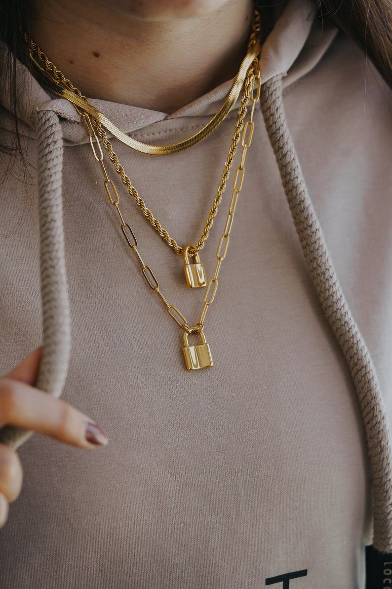 May include: Three gold necklaces with padlock pendants. The necklaces are layered on a person's neck. The top necklace is a thin, flat chain. The middle necklace is a thicker, rope chain. The bottom necklace is a chain with small, rectangular links.
