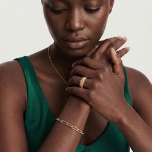 May include: A gold-colored jewelry set featuring a ring, bracelet, and necklace. The ring has a ribbed design, while the bracelet has a chain link style. The necklace is a delicate chain. The jewelry is worn against a green top.