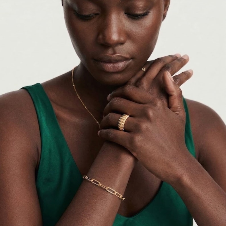 May include: A gold-colored jewelry set featuring a ring, bracelet, and necklace. The ring has a ribbed design, while the bracelet has a chain link style. The necklace is a delicate chain. The jewelry is worn against a green top.