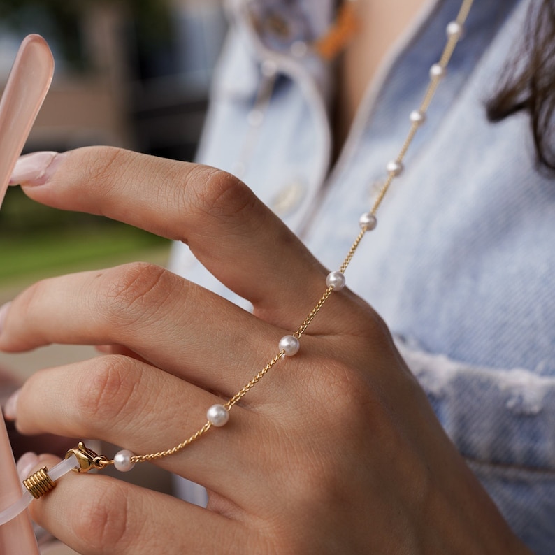 May include: A delicate gold chain with small, evenly spaced pearls. The chain is attached to a pair of eyeglasses. The background is a blurred view of a person wearing a denim jacket.