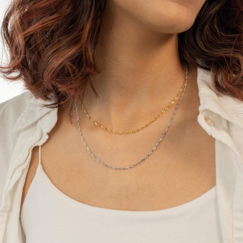 Op de afbeelding: Een vrouw draagt een wit shirt en een gouden en zilveren kettingketting. De ketting is in de stijl van een delicate, gedraaide ketting.